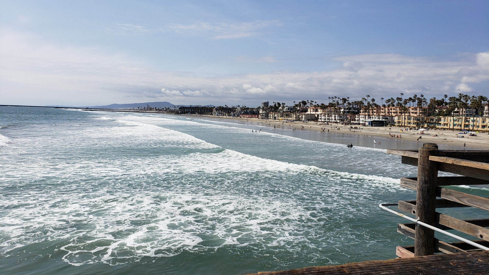 Photo from the Oceanside pier looking out onto the beach and north towards the Oceanside harbor. You can see many palm and a blue sky with clouds in the wayward distance.