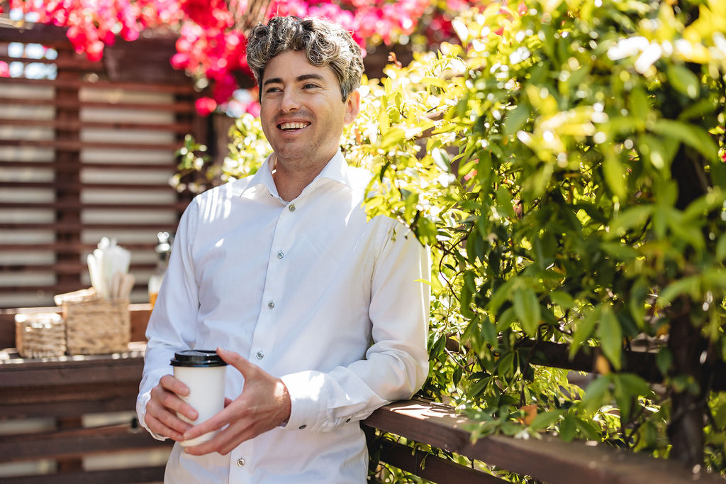 Matthew in a white shirt leaning against a wooden railing, holding a coffee cup, smiles under bright sunlight. Vibrant red and yellow flowers form the background, creating a cheerful atmosphere.