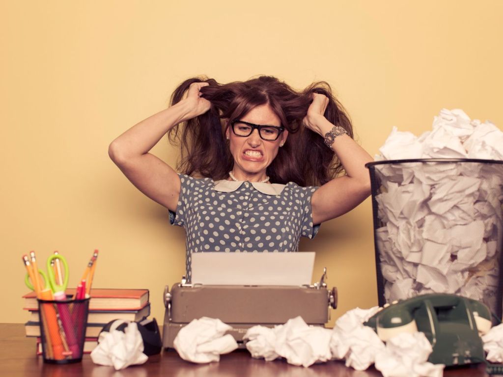 Woman looking frustrated, gritting her teeth and pulling her hair looking at an old pink typewriter sitting at a desk. There are crumpled papers strewn about the desk and a wastebasket full of crumpled paper. Behind her is a yellowish, orange wall.