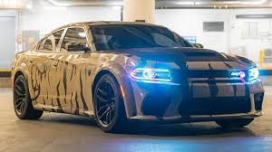 A picture of Miles Hudson's car from social media, the souped-up Dodge Charger called the Hellcat. It has black stripes over a tan body with blue headlights. This photo was taken in a parking garage.