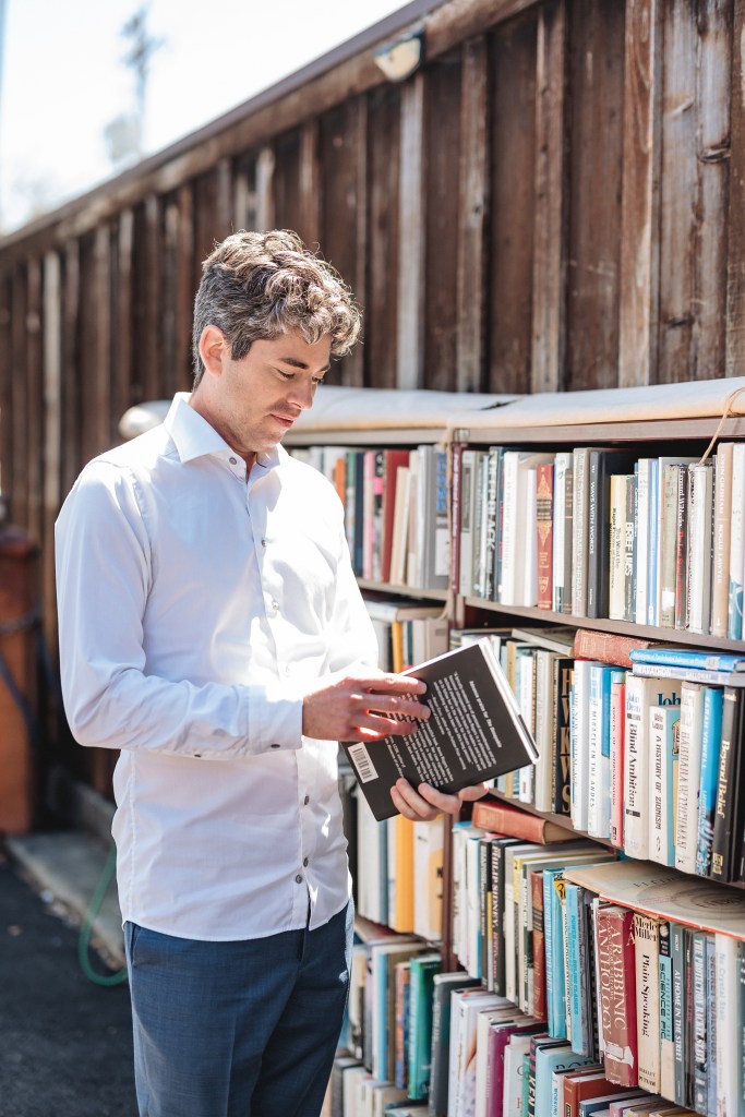 Picture of Matthew outside D.G. Wills bookstore on the right side of the shop looking at a book about to open it at an outdoor bookcase.. He is wearing a white shirt with blue pants.