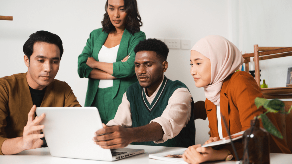 A picture of a diverse group of people all looking at a laptop working together on a school or work project. A man off to the life is holding the computer while another man is typing. A woman stands behind them looking at the screen. Another woman, off to the side and closest to the camera is smiling, doing the same. 