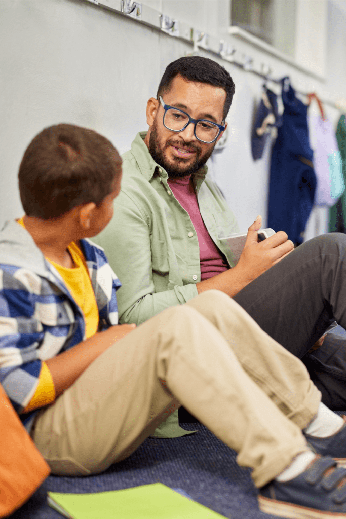 An older man with glasses, a beard, and a green jacket is sitting in a school setting with a young boy, both engaged in a friendly conversation.