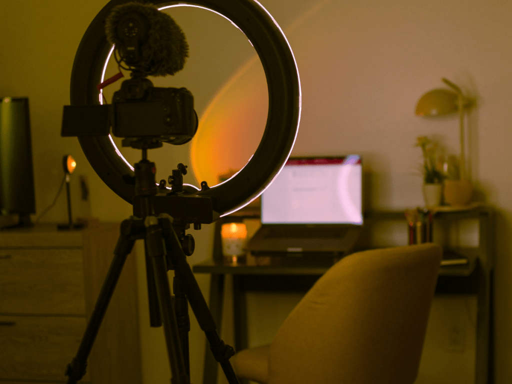 Perfect beginner creator setup: A lit workspace with a camera on a tripod and a ring light in the foreground. A laptop on a desk is open, surrounded by a lamp, plant, and office supplies. A cozy chair is positioned in front of the desk.