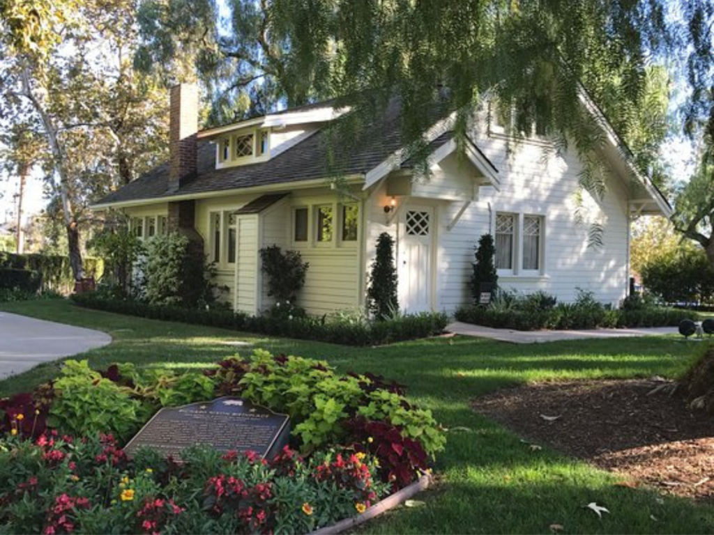 Richard Nixon's Birthplace: A charming white cottage with a sloped roof, surrounded by lush greenery and colorful flowers. A plaque commemorating the site is in the foreground amidst vibrant red and green plants. Trees provide shade, casting dappled sunlight on the scene.