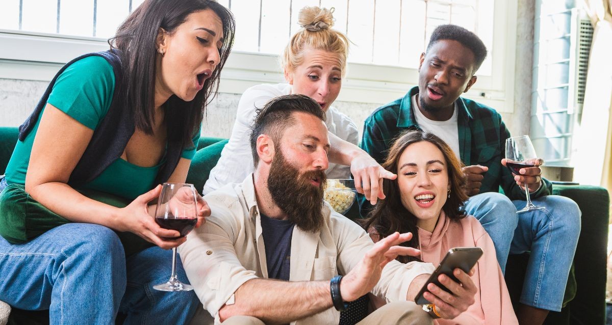 A group of five friends is sitting indoors, looking at a phone one person is holding. They appear excited and engaged. Two of them are holding glasses of red wine, and one is holding a bowl of popcorn. They are seated closely on a couch.