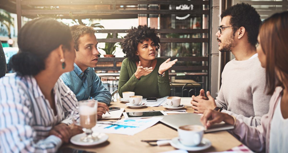 A group of five people sitting at a café table, engaged in discussion. They have cups of coffee and documents on the table. One person is gesturing while speaking, and the others listen attentively. Sunlight filters through the windows.