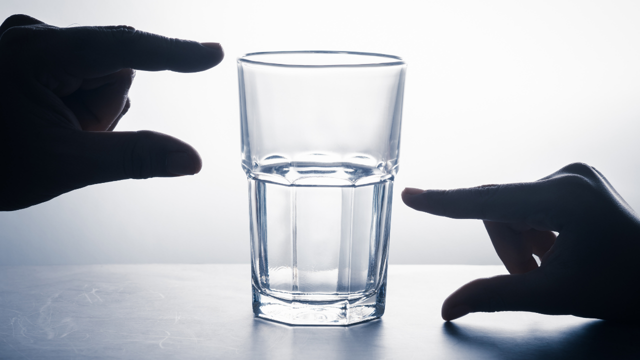 Two hands frame a half-full glass of water against a bright background, creating a silhouette effect. The hands are positioned on either side of the glass, one pointing to the glass half full (optimist) and the other to the glass half empty (pessimist).
