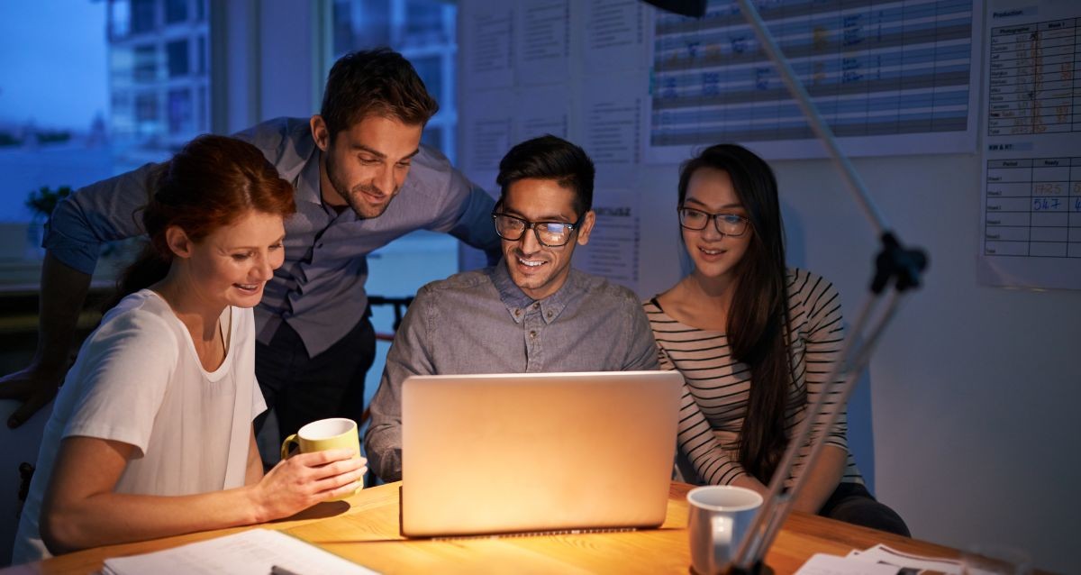A group of four people gather around a laptop at a wooden table, smiling as they work together. Papers and charts are visible on the wall, and a person holds a mug. The room is dimly lit, creating a cozy atmosphere.