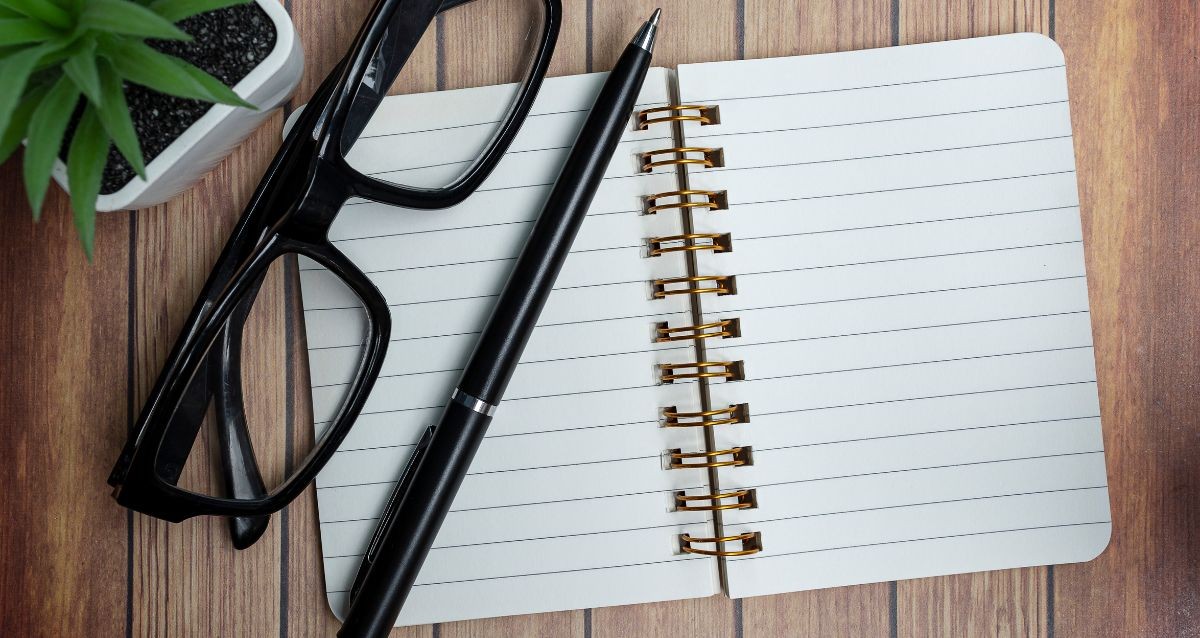 An open lined notebook with a spiral binding, a black pen, and black-framed glasses are laid on a wooden surface next to a small potted plant.