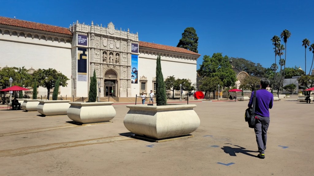 Dr. Matthew Pincus walking in Balboa Park plaza ready to capture cinematic shots of art for local museums. It is a beautiful Southern California day with palm trees and blue sky.